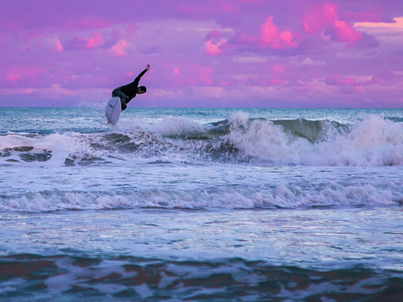 a surfer at sunset catching waves with a pink, blue and purple sky in the background
